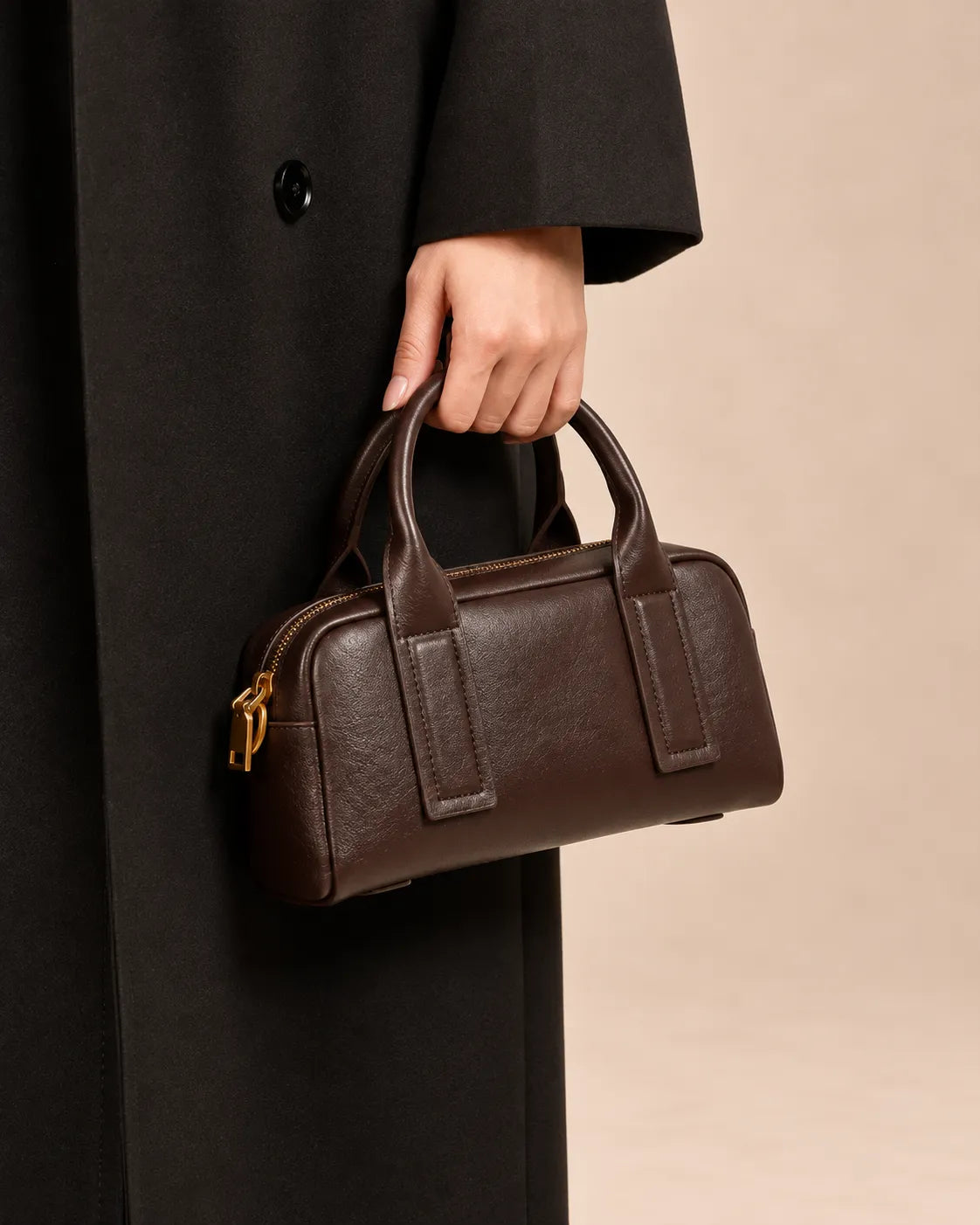 Model holding the Brigitte genuine leather Boston bag in dark chocolate by its top handles, styled with a long black coat, by Malena London