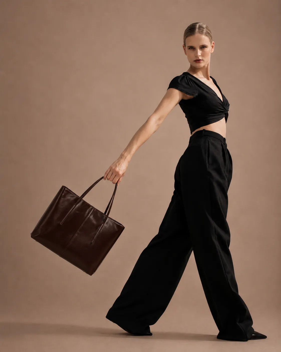 Model carrying the Margaux large leather tote bag in dark chocolate leather while walking in a studio setting