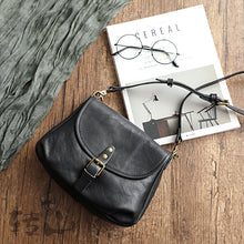Vintage genuine leather shoulder bag in black with a buckled flap and adjustable strap, shown on a wooden table with a magazine and glasses, by Malena London