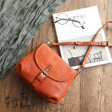 Vintage genuine leather shoulder bag in rust orange with a buckled flap and adjustable strap, shown on a wooden table with a magazine and glasses, by Malena London