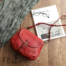 Vintage genuine leather shoulder bag in red with a buckled flap and adjustable strap, shown on a wooden table with a magazine and glasses, by Malena London