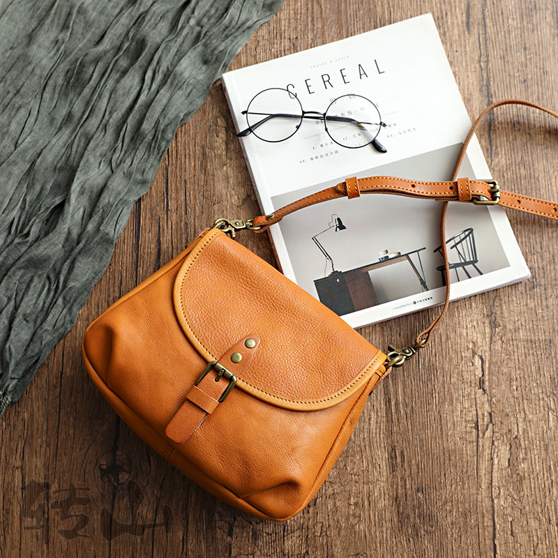 Angled view of the vintage genuine leather shoulder bag in tan with a buckled flap and adjustable strap, shown on a wooden table with a magazine and glasses, by Malena London
