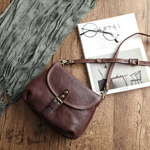 Vintage genuine leather shoulder bag in dark brown with a buckled flap and adjustable strap, shown on a wooden table with a magazine and glasses, by Malena London