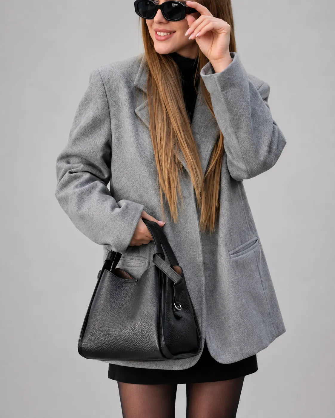 Woman wearing The Amelia black genuine leather handbag styled with a tailored grey blazer and loafers. Structured leather bag for women combining timeless elegance, everyday functionality and contemporary city style.