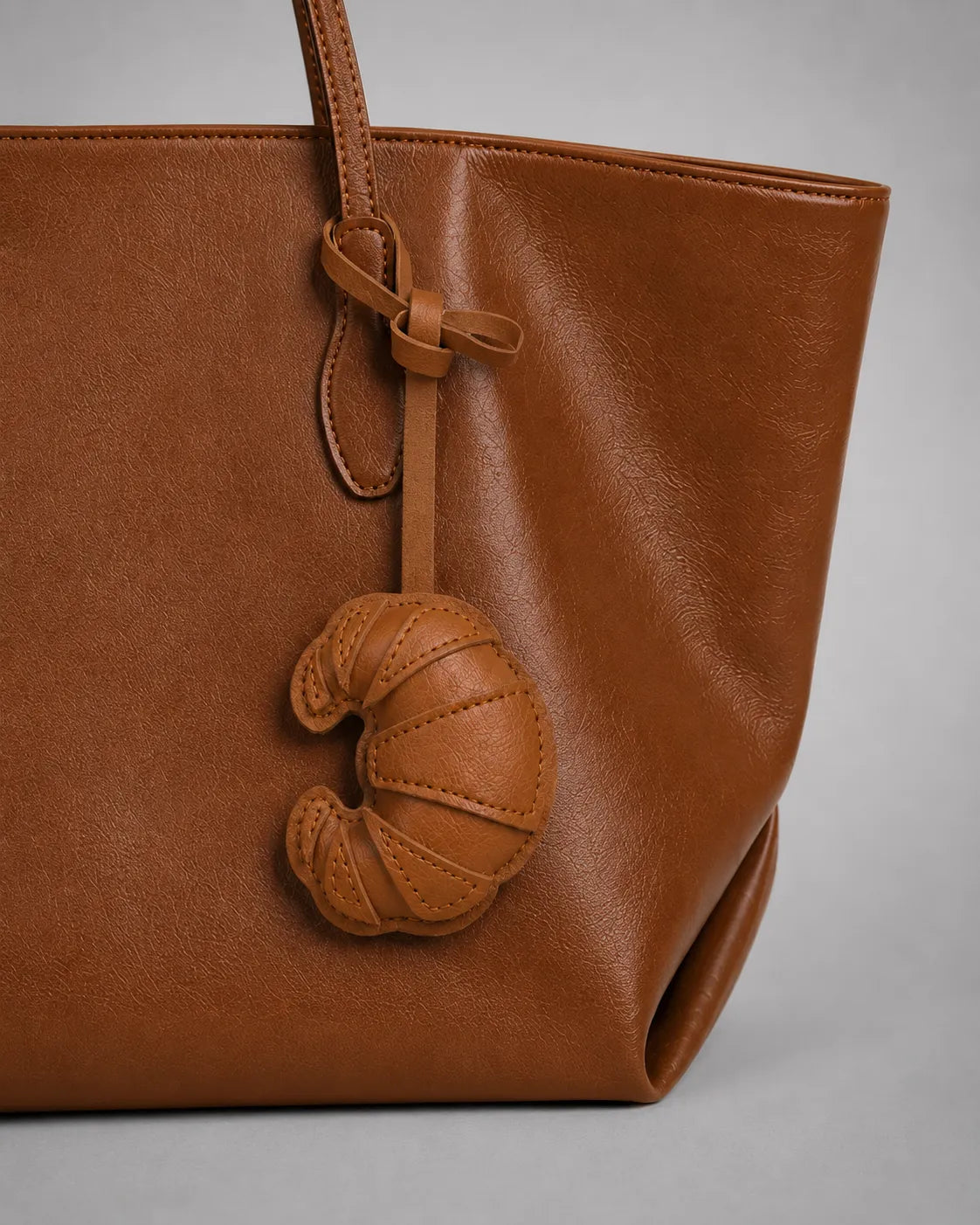 Brown croissant bag charm attached to a tote bag, shown in a close-up view highlighting handbag personalisation and accessory styling.