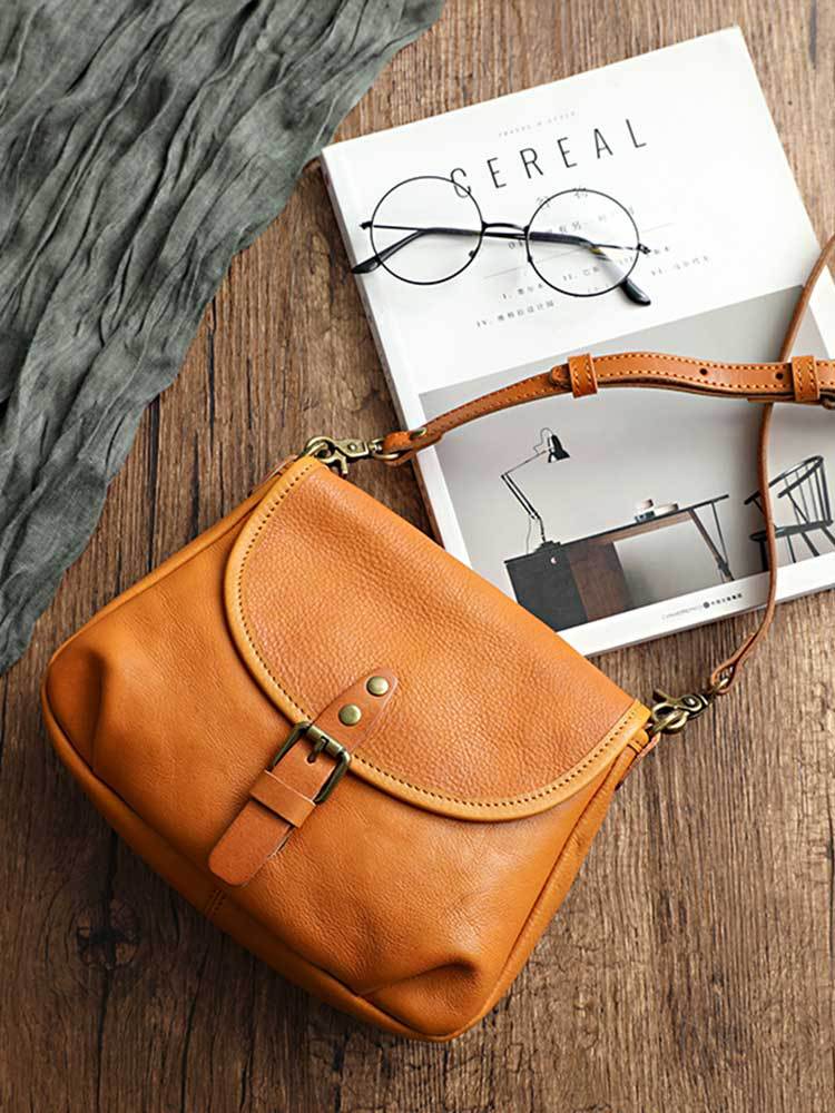 Vintage genuine leather shoulder bag in tan with a buckled flap, brass-tone hardware and an adjustable strap, shown on a wooden table, by Malena London