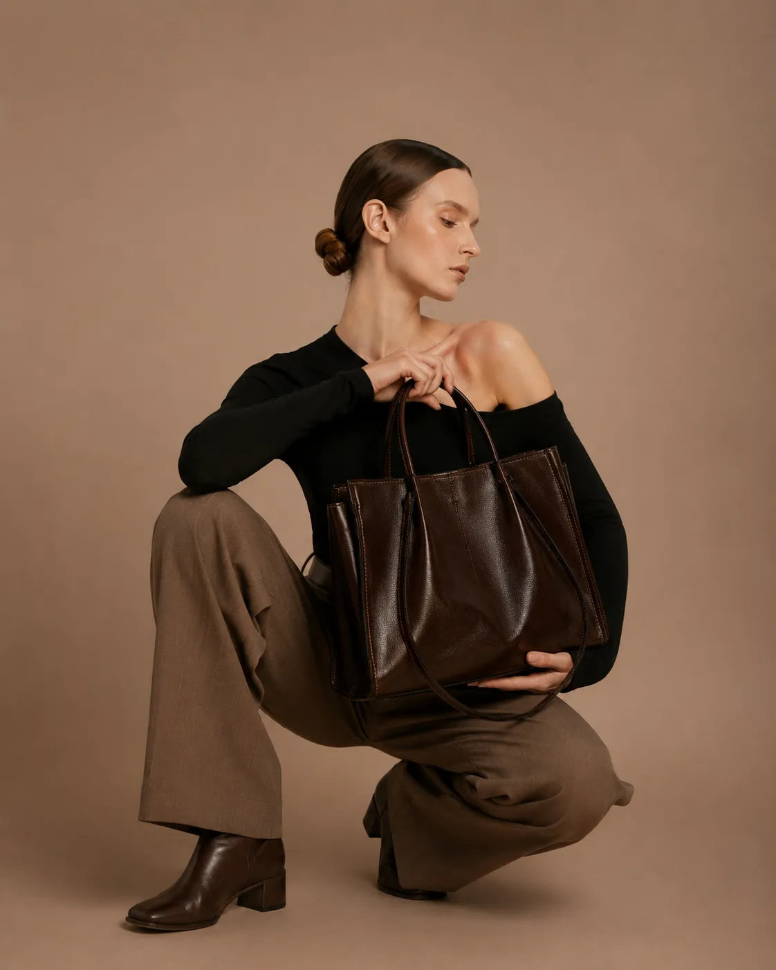 Woman holding the Edith Leather Tote Bag in Dark Chocolate genuine leather, styled with tailored trousers and leather ankle boots against the Malena studio beige background, showcasing the bag’s structured silhouette and elegant city-ready design from The City Edit collection by Malena London.