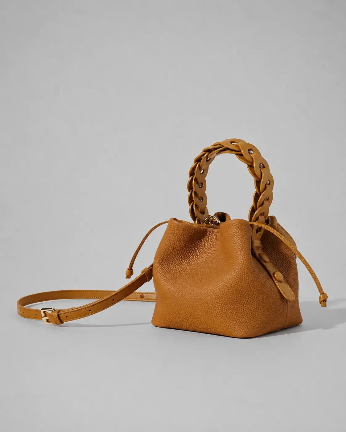 Side angled view of the Léonie genuine leather bucket bag in Brown featuring a removable adjustable shoulder strap, braided leather handle, drawstring closure and modern statement bucket silhouette by Malena London.